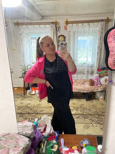 The media file shows a young woman taking a mirror selfie in a room. The room appears to be a bedroom or living area, with a window covered by sheer curtains. The walls are a light color and there is a patterned carpet on the floor. A dresser or shelf is visible to the right of the woman, with various items on it, including bottles, containers, and possibly toiletries. A pink eye mask with black dots is hanging on the wall above the mirror.
The woman is wearing a bright pink zip-up hoodie over a black shirt. She has blonde hair pulled back in a ponytail and is smiling at the camera. She is holding her phone in her right hand, and her left hand is resting on her hip. The image is taken from a slightly low angle, and the lighting suggests it is daytime.
The reflection in the mirror shows more of the room, including a clock on the wall and some bags and items on the shelf. The overall impression is a casual, personal moment captured in a home environment. The text "БАЛДЕЙКА" is visible on a green bottle in the reflection.