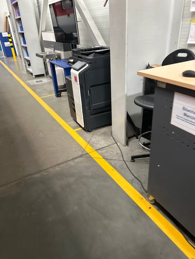 The image depicts a section of an industrial or warehouse-like interior. A prominent yellow line marks a boundary on the grey concrete floor. To the left, tall white shelving units are visible, containing blue binders and other items. In the mid-ground, a black and white printer-copier is positioned on a blue cart. Adjacent to the printer is a sink with a faucet, mounted on a wall. A large screen, possibly a monitor, is mounted above the sink. To the right of the printer, a portion of a light-colored desk is visible, with a black office chair partially obscured. A white sign with blue text is attached to the side of a dark gray cabinet located in the foreground. The environment appears to be well-lit, with visible light fixtures on the ceiling. The overall scene suggests an organized workspace within a commercial or industrial facility, possibly located in Białki, Poland.