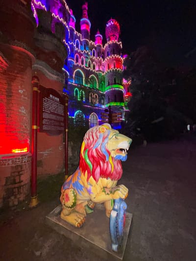 At night, a brightly colored lion statue is in front of a building festooned with multi-colored lights. The lion is lying down, predominantly yellow, with a mane of red, white, and blue. There are green and yellow floral designs on its shoulder. The lion rests on a gray surface, while a blue object is under its paws. Behind the lion, a red brick building is illuminated with red lights. A sign with text in Chinese is visible on the building's surface. The building behind is elaborately decorated with strings of multi-colored lights, forming the shape of castle towers. The background is dark, suggesting it is nighttime. Given the location in Baqiao, China, this appears to be a decorative display at a tourist site or park.