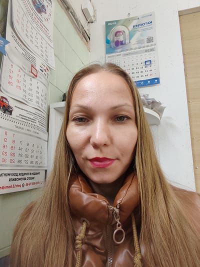 The image shows a medium close-up selfie of a woman with long blonde hair, taken indoors. She has light skin, light green eyes, and is wearing red lipstick. She is wearing a brown puffer jacket. Behind her, on the wall, are several calendars. One calendar, at the top right, is partially visible and features colorful graphics and text. Below it, another calendar is also visible, showing days and dates, with some text in Cyrillic script. Further down, more calendars are visible, some with images of vehicles and rows of dates and text in Cyrillic. The wall appears to be light-colored, and there are some pipes or wires running along it. The lighting suggests it might be daytime, but it's difficult to determine precise time or weather conditions. The woman's expression is neutral. The calendars suggest a domestic or office setting, possibly related to automotive services given the vehicle images. The visible text is primarily in Cyrillic, with some numbers and what appear to be dates.