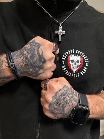 The scene depicts a close-up of an individual's upper torso and hands, situated in the city of Grevenmacher, Luxembourg. The subject, a male, is wearing a black zippered jacket. On the left chest area, a white embroidered circular logo reads "SUPPORT CRUZADOS 2012 MOTORCYCLE CLUB" encircling a white skull with red accents. A silver-colored chain necklace with a textured Maltese cross pendant, featuring a red and white central emblem, hangs from the neck.
Both of the subject's hands are clenched into fists and are heavily tattooed with intricate grey-scale designs across the knuckles and dorsum. The right hand (viewer's left) wears a multi-strand black braided leather bracelet adorned with four metallic cylindrical beads engraved with names: "Sheyenne," "Shalina," "Abby," and a fourth partially obscured name. A silver-colored ring with a red central emblem is present on the right middle finger, and another dark-colored ring is on the right ring finger. The left hand (viewer's right) displays a dark grey or black ring on the ring finger and a black smartwatch with a black strap on the wrist. The background behind the subject is indistinct and dark.
