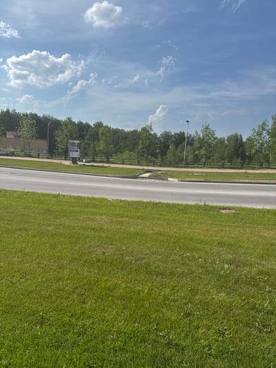 The image shows a bright, sunny day in a semi-urban or park-like setting in Koltsovo, Russia. A paved road runs horizontally across the middle of the frame, with a well-maintained green lawn in the foreground. Beyond the road, a sidewalk is bordered by a wooden fence, and behind that, a dense line of deciduous trees forms a backdrop. A tall lamppost stands near the fence, and a modern building with a light-colored facade and a dark roof is visible on the far left. A tall, rectangular advertisement stand is positioned near the building. The sky is a vibrant blue with scattered white cumulus clouds. There are no people visible in the scene. The environment suggests a public space, possibly a park or a street leading to a recreational area.