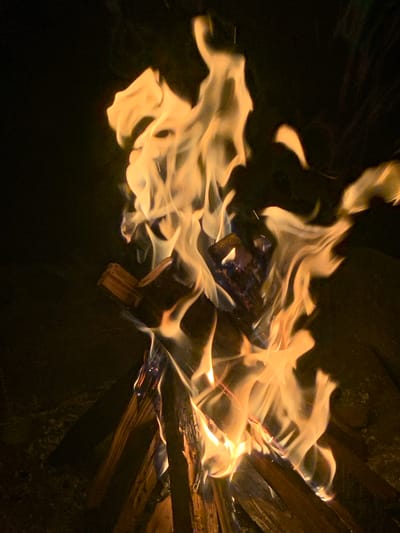This is a close-up, low-light photograph of a campfire in Tabagrebi, Georgia. The fire is burning brightly, with yellow and orange flames licking upwards and consuming several pieces of wood. The wood is arranged in a rough pile, with some pieces appearing charred and others still intact. The background is dark and out of focus, suggesting the photograph was taken at night. There are no people or animals visible in the frame. The overall impression is one of warmth and primal energy, with the dancing flames dominating the scene.