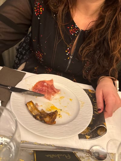 A partial view of a woman at a dining table. She has long brown hair and wears a black top with a zippered front and patterned design. On the table is a white plate with remnants of a meal: ham, a bone, and sauce. A knife rests on the plate. A dark napkin and a spoon are beside the plate. A name card is visible, and she wears a smartwatch on her wrist.