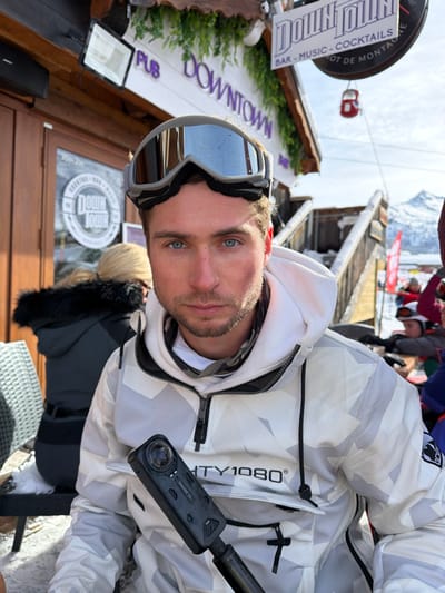 The image displays a Caucasian male subject seated outdoors in a snowy environment in front of the "Downtown" establishment, located in Les Allues, France. The male, with blue eyes and light facial stubble, wears a white and grey geometric camouflage-patterned anorak-style ski jacket with "MIGHTY1080®" branding on the chest. Grey mirrored ski goggles are positioned on his forehead. He holds a black Insta360 X4 camera on a black extension pole in his right hand, oriented towards his left side, and directly faces the primary camera. To the subject's left, partially obscured, is another individual with blonde hair, wearing a dark jacket with a black fur-lined hood, seated at an outdoor table. The background features a wooden building with a sign displaying "PUB DOWNTOWN," "BAR - MUSIC - COCKTAILS," and "SPOT DE MONTAGNE," embellished with artificial green foliage. Snowy wooden steps are visible to the right of the building. In the far background, a red gondola cabin is suspended, and snow-capped mountains rise under a clear blue sky. Snow is present on the outdoor seating and ground surfaces.