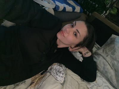 A young person is lying on a bed in a dimly lit indoor setting, gazing directly at the camera. The individual, who appears to be female, has dark hair, strong eyebrows, and a visible nose piercing. They are dressed in a dark, long-sleeved top or hoodie, and a small, illegible tattoo is noticeable on their left wrist or forearm.
The bed features patterned bedding, with one pillow displaying various textual elements. Partially discernible French phrases such as "est curieuse. Pas seulement" and "à grands" are visible on the fabric, along with "me style, autrefowances a". Other less coherent text segments like "EL E" and "[ ΦΕΥ" are also present. In the bottom left corner, some green leafy plants and a wooden surface provide a glimpse of the room's decor. A blue and white striped fabric item is also partially visible on the bed near the person.
The low lighting conditions suggest that the image was taken during the evening or night. The person's expression is neutral and relaxed, indicating a casual moment, possibly a selfie or a close-up portrait. The scene depicts a personal space, likely a bedroom, situated in Belopoptsi, Bulgaria.