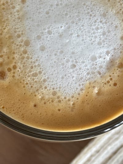 This is a close-up, top-down shot of a beverage with a frothy top layer. The beverage appears to be coffee, with a gradient from a creamy brown at the bottom to a lighter, white foam at the top, dotted with small bubbles. The edge of a clear glass is visible at the bottom of the frame, resting on a textured surface that is likely a table or placemat. The background is out of focus and suggests an indoor setting. There are no people or specific landmarks identifiable in the image, and therefore, no location in the city of Гамово, Russia can be determined.