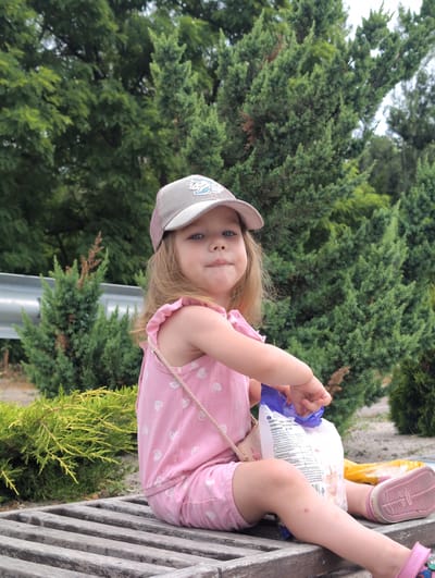 In Perliavka, Ukraine, a young girl wearing a pink dress with white hearts and a gray baseball cap sits on a wooden bench. She is holding a white bag with blue accents, seemingly trying to open it. The background is filled with lush green trees and bushes, suggesting an outdoor setting, possibly a park or natural area.