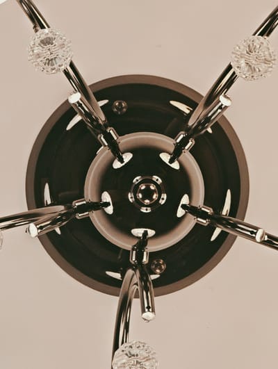This is a close-up, overhead shot of a modern chandelier. The chandelier has a dark, circular base from which several polished metal arms extend outwards. Each arm is tipped with a clear, faceted crystal orb, catching the light. The central part of the chandelier features a layered design with a polished metal fixture. The background is a plain, light-colored surface, likely a ceiling. The lighting appears to be ambient, casting soft reflections on the metallic surfaces. The image is a still shot, with no activity or event taking place. The focus is entirely on the intricate details of the chandelier's design. There is no visible text in the image.