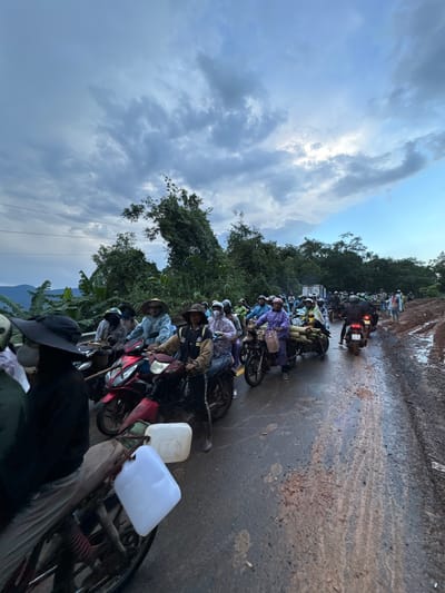 A long line of motorcyclists, many wearing conical hats and raincoats, are stopped on a wet, muddy road. The road appears to be undergoing some sort of work or has been affected by recent rain, with significant mud and debris. The motorcyclists are a mix of individuals, some appearing to be carrying goods such as bamboo poles. In the background, lush green foliage and trees suggest a rural or mountainous area. The sky is overcast with dramatic clouds, and the overall lighting indicates it is either early morning or late afternoon. The scene appears to be in the vicinity of Nao Sẻ, Vietnam, given the context.