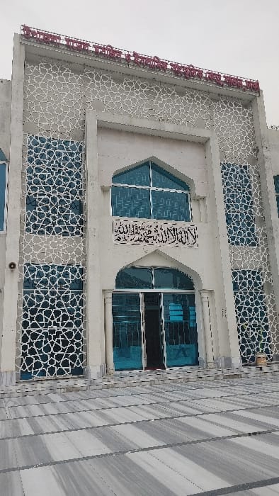 The image displays the front facade of the Upazila Social and Cultural Center, situated in Savar, Bangladesh. The building is a light-colored, multi-story structure with a modern architectural style incorporating Islamic design elements. Its exterior features extensive vertical sections of white geometric lattice patterns overlying or integrated with large, blue-tinted glass windows. A central, recessed arched entryway contains partially open double blue-tinted doors, providing a glimpse into the dark interior. Above this entrance, a horizontal panel displays black Arabic calligraphy, which translates to the Shahada ("There is no god but Allah, Muhammad is His Messenger"). A large, arched, multi-pane window is positioned above the calligraphy. Along the top edge of the building, red Bengali script identifies the structure as an "Upazila Social and Cultural Center." The foreground consists of a meticulously paved plaza composed of large, rectangular tiles with alternating light gray and white stripes, laid horizontally, leading directly to the building's entrance. The sky overhead is overcast. No individuals, vehicles, or active scenes are present within the frame.