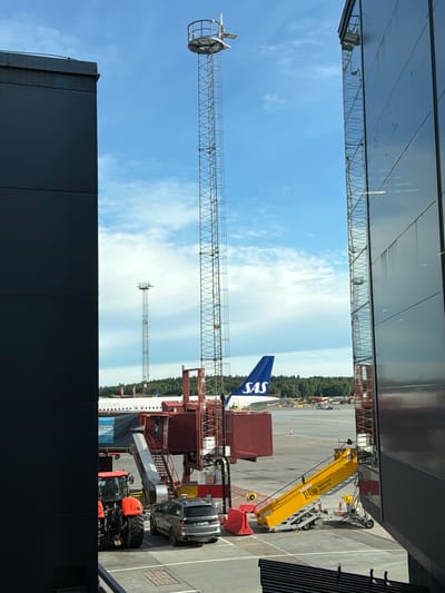 The image depicts an airport scene at Stockholm-Arlanda Airport in Sweden. A SAS airplane is parked at a gate, connected by a jet bridge. A red tractor is positioned near the jet bridge, and a silver SUV is parked close by. In the foreground, a yellow mobile stairway with the Swedavia Airports logo is set up next to the airplane. Two tall communication towers are visible in the background, along with a wooded area and a blue sky with scattered clouds. The scene is framed by the dark, modern architecture of the airport terminal buildings.