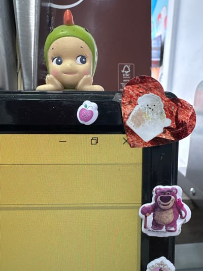 The image displays a section of what appears to be an electronic device screen. The screen's surface is yellow with vertical lines. Affixed to the device's black frame are various stickers and a toy. Stickers include a red heart with a white blob, a pink bear, and a pink flower. A small, green-helmeted doll peeks out from behind the frame's edge. A brown box is partially visible in the background.