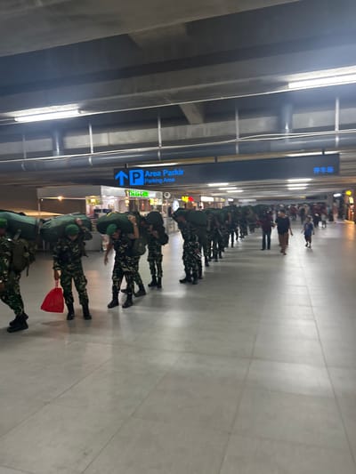 In Baji Mangngai, Indonesia, soldiers wearing camouflage uniforms and green helmets are seen marching in a line indoors, carrying large green duffel bags on their shoulders. The first soldier from the left is also holding a red bag. The soldiers appear to be in an airport or a large public building with a tiled floor. Above them, there are signs with directional arrows, one indicating "Area Parkir Parking Area" in blue and white. Another sign is visible in the distance with what appears to be Chinese characters. The lighting suggests it is nighttime or an indoor environment with artificial lighting. Other individuals, including civilians, are visible in the background, walking in the same direction as the soldiers. The environment is clean and well-lit.