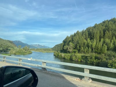 The image captures a scenic view of a wide, calm river flowing through a valley, as seen from a bridge. The left side of the frame shows a mountainous landscape with green slopes and distant peaks under a partly cloudy blue sky. The river curves to the right, with lush green vegetation and trees lining its banks. A dense forest of evergreen trees covers the hillside on the right side of the image, extending up towards the sky. In the foreground, a section of the bridge's concrete abutment and metal railing is visible, with a car's side mirror reflecting the road and surrounding scenery on the lower left. The overall scene suggests a natural, tranquil environment, likely in a rural or mountainous region. The image was taken from a moving vehicle on the bridge.