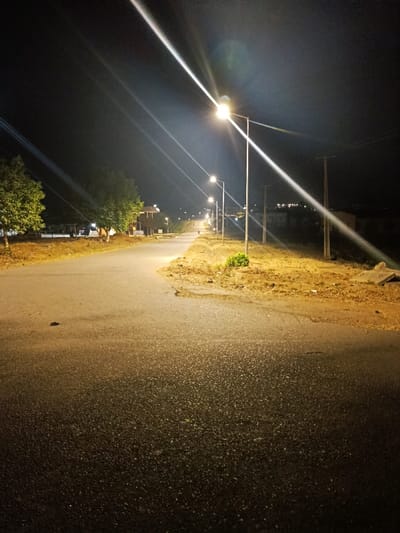 The image depicts a deserted asphalt road at night in Rafin Pa, Nigeria. The road extends into the distance, curving gently to the left in the mid-ground. A small, dark, unidentifiable object lies on the asphalt in the lower-left foreground. Multiple streetlights on tall poles line the right side of the road, casting bright, warm light; the closest streetlight exhibits pronounced lens flares. The asphalt surface appears textured, reflecting the illumination more intensely towards the mid-ground. To the left of the road, green-leaved trees are present, with partially visible, indistinct structures behind them. The ground on both sides of the road consists of dry earth with sparse, low vegetation. Wooden utility poles carrying overhead lines are visible further down the right side, alongside the streetlights. Distant structures are faintly discernible against the dark sky in the background. No individuals, animals, or vehicles are present.