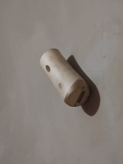 A small, cylindrical, light-brown wooden object sits diagonally against a mottled, pale-grey background. The object has two small, dark, circular holes on its side. One end has a darker-brown cap-like structure with a rectangular slot in its center. A distinct shadow of the object is cast on the background.