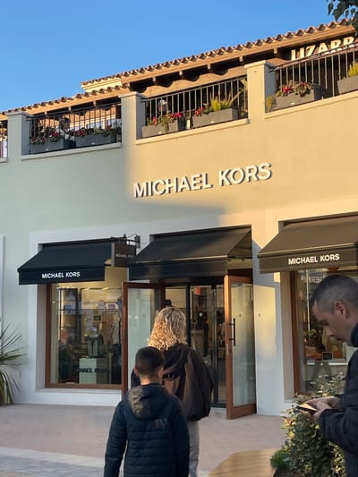The image displays an outdoor scene at the Mallorca Fashion Outlet in Marratxí, Spain. The primary subject is the storefront of a Michael Kors store, identifiable by the large white "MICHAEL KORS" lettering on its light cream-colored facade. The store features multiple black awnings over its display windows and a double glass door entrance with brown wooden frames, which is open. Merchandise is visible through the store's glass front.
Above the Michael Kors store, a second story features a balcony with black wrought-iron railings and grey planter boxes containing green foliage and red flowers. A partial sign for "LIZARD" is visible on the upper level, to the right, behind the balcony railing. Several individuals are present on this balcony.
In the foreground, an adolescent male with short brown hair, wearing a dark blue puffer jacket, stands with his back to the camera. Immediately in front of him, a female with curly blonde hair, wearing a dark jacket, is also seen from behind, positioned near the entrance of the Michael Kors store. To the right, partially obscured by low green shrubbery, a male with short dark hair is visible in profile, looking down at a smartphone held in his hands. The ground is a light-colored paved walkway. The scene is illuminated by natural sunlight, casting soft shadows, and a clear blue sky is visible at the top.