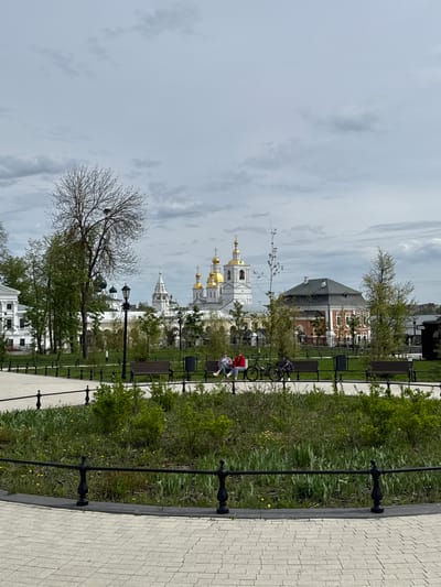 The image captures a view of Arzamas, Russia, on an overcast day. It features a paved walkway bordered by a low black metal fence and green vegetation. Further beyond, there's a park area with benches, a bicycle, and people sitting, seemingly relaxing. In the background, there is a complex of buildings, including the Cathedral of the Resurrection with its golden domes, indicating its religious significance. There are also other structures with varying architectural styles, including one that appears to be a traditional brick building. The sky is filled with clouds, contributing to a somber atmosphere. A bare tree stands in the foreground.