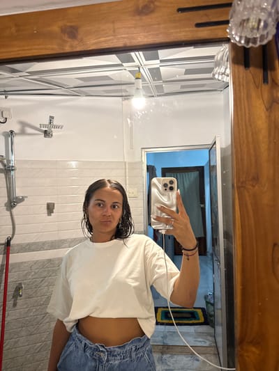 An adult female is depicted taking a mirror selfie in a bathroom located in the city of Nilwella, Sri Lanka. She possesses wet, dark hair and a medium complexion. Her attire consists of a white short-sleeved crop top and light blue high-waisted denim jeans. She wears a ring on her left ring finger and a dark bracelet on her left wrist. Her facial expression includes wide-open eyes and pursed lips.
The subject holds a light-colored smartphone with a ridged case in her left hand, extended towards the mirror. A white charging cable is connected to the phone and extends downwards. The mirror, framed by dark wood, reflects a bathroom environment. The walls are tiled in white with a horizontal band of darker tiles. A chrome shower head and a faucet are mounted on the left wall. A red-handled object, possibly a brush or mop, leans against the wall on the far left.
The ceiling features white square panels with a black and white checkered pattern. A bare incandescent light bulb hangs centrally from the ceiling, illuminated. A decorative clear glass light fixture is partially visible on the upper right side of the mirror frame. Through an open doorway reflected in the mirror, a room with light blue walls and a darker door is visible. A small, patterned mat lies on the floor in the reflection of this doorway.