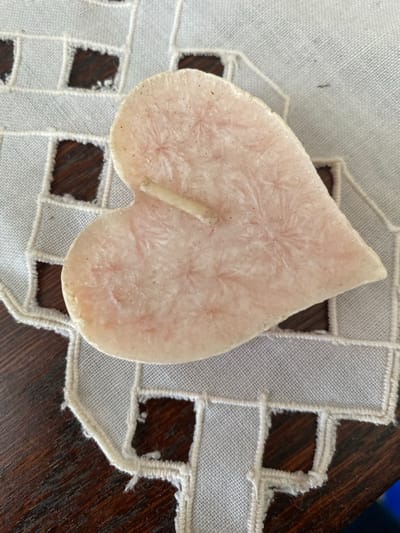 A close-up, high-angle shot focuses on a light pink, heart-shaped candle with a rough, crystalline texture and a small wick protruding from its center. The candle rests on a white embroidered doily with square cutouts that reveal the dark wood grain of a table beneath. The doily's edges appear slightly frayed. The scene is static and appears to be indoors, likely in a home setting. There are no people or animals present, and no specific landmarks are visible to indicate a precise location within Osek, Czechia.