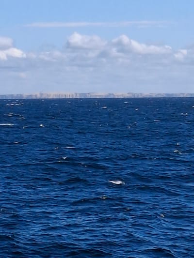 The image shows a body of water, presumably the sea, with a clear blue sky and some clouds visible in the background. The water is a deep blue color with ripples and waves creating texture on the surface. There are some whitecaps or small splashes of water visible. In the distance, there might be a coastline or landmass but it is not clearly defined. The location is Unknown, Denmark.
