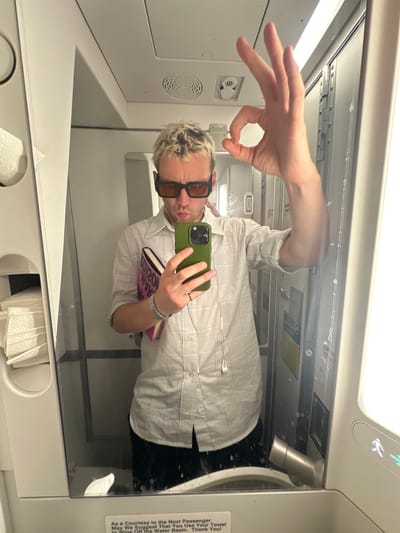This is a self-portrait photograph taken in a mirror, likely on an airplane or similar mode of transport. The subject, a person with blonde hair and sunglasses, is holding a green smartphone and a pink book. They are wearing a light-colored, collared shirt with subtle grid patterns.
The background, visible in the mirror's reflection, shows the interior of a cabin, with overhead compartments and lighting. There are also signs with what appear to be safety or courtesy instructions. One sign reads: "As a Courtesy to the Next Passenger May We Suggest That You Use Your Towel to Wipe Off the Water Basin. Thank You!".
The image is taken from a low angle, and the person is making a gesture with their hand, forming a circle with their thumb and index finger. The overall mood appears casual and somewhat playful. The lighting suggests it is daytime, or at least well-lit within the cabin. No specific weather conditions are discernible. The location context provided is Luton, United Kingdom, which could imply the person is traveling to or from this location.
