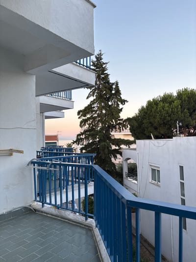 This media file captures an outdoor scene in Nea Irakleitsa, Greece, likely during the late afternoon or early evening, as suggested by the soft, diffused light and the gradient of colors in the sky. The perspective is from a slightly elevated viewpoint, looking out towards a building with distinct architectural features.
The primary subjects are the architecture and the surrounding environment. We see elements of a white, modern building with a sharp, angular design, possibly part of a residential complex or hotel, featuring triangular roof-like structures and a blue metal staircase or balcony railing. Behind this, the branches of a tall, mature evergreen tree dominate the upper left and center of the frame, its needles creating a textured canopy against the pale sky. Further in the distance, other buildings are visible, some with red-tiled roofs and windows, hinting at a Mediterranean or southern European setting. The ground level shows patches of greenery and what appears to be a gray tiled wall.
There are no people clearly visible in the image, nor is there any obvious activity or event taking place. The focus is on the static elements of the scene – the structures, the vegetation, and the sky.
Notable details include the stark contrast between the white geometric forms of the building and the natural green of the tree. The blue of the railing adds a vibrant accent. The sky exhibits a subtle transition from a clear, light blue at the top to softer hues of pink and yellow closer to the horizon, indicating either sunset or sunrise. The overall impression is one of tranquility and understated modernity within a natural setting. No visible text is present in the image.