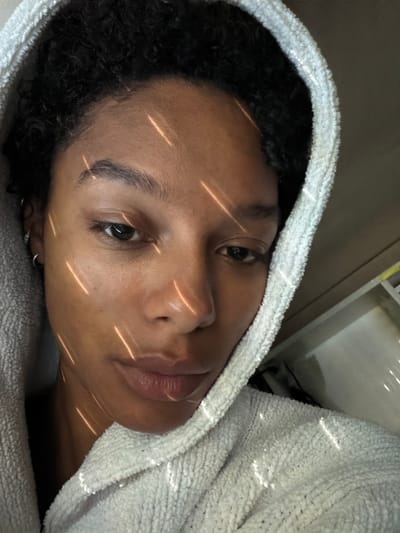 A young adult female, appearing to be of mixed or Black ethnicity, is depicted in a close-up, slightly downward-angled portrait. She has dark, curly hair visible beneath a light-colored, likely white or off-white, terry cloth hooded garment, which frames her face and shoulders. Her skin tone is medium brown, and she has dark eyes with a relaxed, slightly pensive expression. Small silver hoop earrings are visible on her left ear (viewer's right). The scene is indoors, characterized by several distinct, golden-orange diagonal streaks of light cast across her face and the textured fabric of her garment, suggesting natural light filtering through a patterned obstruction like blinds or a window grille. The background is indistinct and out of focus, revealing only muted tones of brown and grey, providing no specific identifying features of a room or exact location. There are no visual indicators within the image to suggest its specific geographic location in the city of Champs-sur-Marne, France, or any other region.