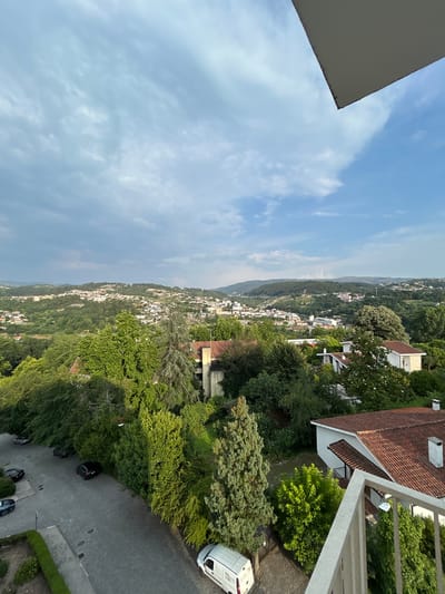 The image shows a panoramic view of Amarante, Portugal, from an elevated perspective. The scene is dominated by a sprawling cityscape nestled within rolling green hills. Numerous houses with red-tiled roofs are scattered across the landscape, interspersed with lush trees and vegetation. A paved street with several parked cars is visible in the foreground, leading towards the town. The sky is partly cloudy with a mix of blue sky and grey clouds, suggesting a dynamic weather pattern. A white van is parked on the side of the street. In the immediate foreground, a balcony railing is visible, indicating the vantage point from which the photograph was taken. The overall impression is one of a tranquil, verdant urban environment.