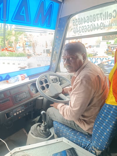 A middle-aged man with a mustache sits in the driver's seat of a bus, looking directly at the camera. He is wearing a light pink collared shirt and gray pants. The bus interior is visible, including the steering wheel, dashboard with instruments, gear stick, and patterned blue and yellow seats. A bottle of water is on the dashboard, and a blue sign with large white lettering is above the windshield. Outside the bus, a street scene is visible with trees, buildings, and other vehicles, suggesting the bus is in transit within the city of Kurnool, India. The man appears to be a bus driver.