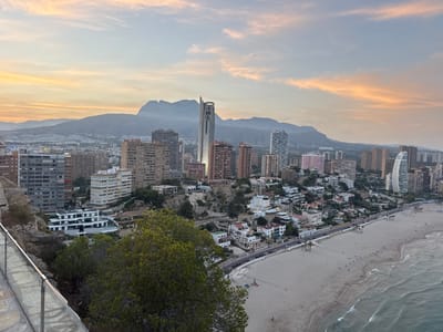 This is an aerial, high-angle outdoor shot of a coastal city in Benidorm, Spain, at dusk. The scene features a sprawling cityscape with numerous apartment buildings and hotels, many of which are tall and modern. In the background, a mountain range is visible under a sky streaked with soft orange and pink clouds, suggesting the sunset or sunrise.

The foreground is dominated by a lush green tree and a rocky cliff face. A modern glass and metal railing system runs along the edge of the cliff, indicating a viewing area or walkway. Below the cliff, the city extends towards a wide, sandy beach. The beach is lined with palm trees and a promenade, where a few cars are parked and a volleyball net is set up. The sea, with gentle waves, meets the shore, and a few people can be seen on the beach and in the water, enjoying the twilight.

The overall mood is serene and picturesque, capturing the beauty of a coastal city transitioning from day to night. The lighting suggests late afternoon or early evening, with the warm tones in the sky creating a peaceful atmosphere. No visible text is present in the image.