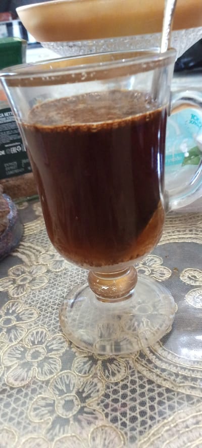 A close-up, eye-level, indoor shot captures a clear glass mug with a stemmed base, filled with dark brown coffee. The coffee has a frothy layer on top and some sediment settled at the bottom. The mug sits on a table covered with a white lace tablecloth featuring a floral pattern. In the background, out of focus, is a light brown bowl or dish. There are no visible people in the image. The scene is static and well-lit, with no indication of Tbilisskaya, Russia.