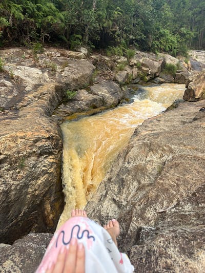 A person's bare feet, adorned with a light-colored garment patterned with pink and blue wavy lines, rest on the edge of a rocky outcrop overlooking a powerful waterfall. The water, a muddy yellow-brown, cascades forcefully down a narrow channel between rough, grey and tan rocks. Lush green ferns and trees form a dense backdrop behind the rocky terrain, suggesting a natural, possibly forested, environment. The scene appears to be a remote outdoor location, likely a natural attraction within the vicinity of Păng Tiêng 1, Vietnam.