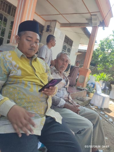 The image captures a casual outdoor gathering, likely on a porch or veranda, under bright daylight. In the foreground, two men are seated.

On the left, a man of East Asian descent, appearing middle-aged, is seated, wearing a black peci (a traditional cap often worn by Muslim men in Southeast Asia) and a long-sleeved yellow shirt with a light blue and white batik-like pattern. His eyes are closed or looking down, and he is holding a smartphone with a purple case in his right hand. His left hand is resting on his thigh. He is wearing dark trousers or jeans.

To his right, an older man, also of East Asian descent, is seated. He has a beard and short, grey hair. He is wearing a patterned long-sleeved shirt in shades of brown and white, and light-colored trousers, possibly khaki or beige. His gaze is directed towards the viewer, with a neutral expression. His hands are clasped or resting on his lap.

In the mid-ground, behind the two men, another man wearing a light-colored t-shirt is visible, looking to the right. Further in the background, a fourth person, partially obscured, can be seen sitting on a piece of furniture, and a fifth person is visible behind the older man's shoulder.

The setting appears to be the exterior of a house. The porch has orange-colored pillars with white vertical stripes and a white ceiling. There are windows with grilles on the house facade. The ground is dirt, suggesting an unpaved outdoor area. Several white plastic chairs, some draped with white cloth, are scattered in the background, along with some plastic bottles and other debris on the ground. Green foliage and trees are visible further in the background on the right, indicating an outdoor, possibly rural or semi-urban environment. The bright lighting suggests it is daytime, likely mid-morning or afternoon, with direct sunlight casting shadows.

A digital timestamp "05/02/2026 14:35" is visible in the bottom right corner of the image.