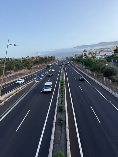 This is a medium-angle, high-contrast, full shot of a multi-lane highway in Adeje, Spain, under a hazy, overcast sky. The highway runs diagonally from the bottom center towards the top right of the frame, with a concrete median dividing opposing traffic. The asphalt is dark and wet, with white lane markings and a textured rumble strip on the edges.
On the left side of the highway, several cars are traveling in the same direction: a white hatchback, a white van, and a green bus. There are also trees, rocks, and some vegetation bordering this side of the road, with buildings and more trees visible in the distance.
On the right side of the highway, a black SUV is visible, along with several other cars further down the road. This side also features vegetation and buildings in the background. The mountains in the distance are partially obscured by fog or clouds. Streetlights are positioned along the highway on both sides. The scene depicts a typical day on a major road, with vehicles in transit and the surrounding urban and natural landscape.