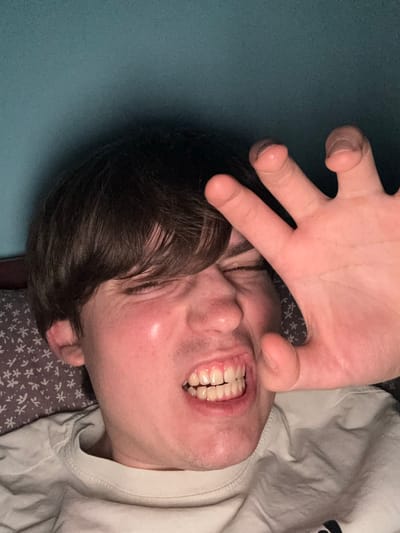 The image shows a close-up of a young person's face, with their right hand raised to cover part of their face. The person has brown hair and is wearing a white t-shirt. Their eyes are closed, and their teeth are clenched. The background is a blue wall. The image is taken from a low angle. There is no information in the image to indicate the location in the city of Dnipro, Ukraine.