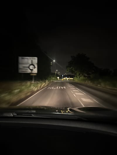 A nighttime scene unfolds from the perspective of a vehicle moving along a road, indicated by a slight motion blur. The image is predominantly dark, with artificial light sources illuminating key features. On the immediate right, a dark section of a car's interior or door frame is visible, reflecting glints of light from outside.
The asphalt road surface is marked with white paint, showing diagonal chevrons that suggest an approach to a junction or a filter lane. The word "SLOW" is distinctly painted in large white letters on the road, oriented for oncoming traffic.
To the left of the road, a narrow grassy verge transitions into a darker area lined with trees, visible as silhouettes against the faint ambient light. Several streetlights are spaced along the roadside, casting light forward and illuminating parts of the road and surrounding vegetation. In the foreground on the left, a prominent white road sign is visible. It clearly displays a black circular arrow symbol, indicating a roundabout or mini-roundabout ahead. Above this symbol, the road number "A41" can be discerned, while other text on the sign, likely directional information, is too blurry to read. Further in the distance along the left, additional streetlights and yellow and black chevron hazard signs are faintly visible.
The scene captures the experience of night driving on a road, with reliance on vehicle headlights and roadside lighting for navigation. The overall environment is dark, suggesting late evening or early morning hours. The road markings and sign types are consistent with those found in the United Kingdom, aligning with the Hertsmere location context.