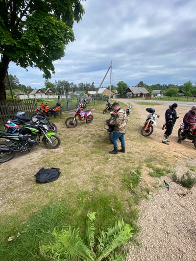 A group of people are gathered with several motorcycles in Парэцкі сельскі Савет, Belarus. One man is bent over a motorcycle, seemingly working on it. Other individuals stand near their bikes, observing. The environment consists of a grassy area with patches of gravel and dirt. There are houses in the background. A tree stands to the left.