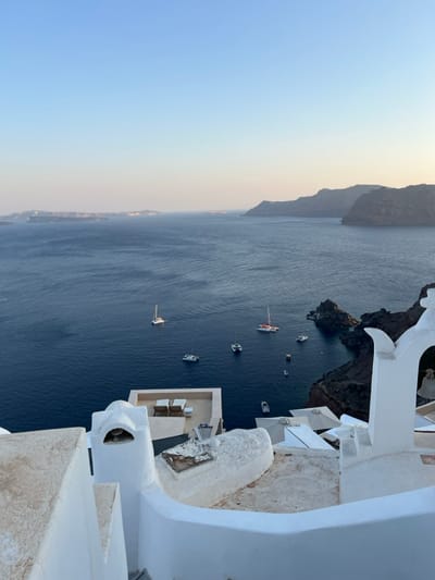 A high-angle, panoramic view overlooks the Aegean Sea and the Santorini caldera from Oia, within the Ia Municipal Unit, Greece. In the foreground, traditional whitewashed Cycladic architecture includes curved walls, staircases, and a flat rooftop terrace with two light-colored lounge chairs. To the right, a distinctive white archway with a visible hanging bell is present, alongside several folded patio umbrellas. The deep blue caldera water in the mid-ground contains approximately a dozen stationary vessels, including several catamarans (one with a white mast, another with red elements) and various smaller motorboats. The background features steep, dark volcanic cliffs to the right, forming part of the caldera rim, and distant, flatter landmasses on the left horizon. The sky is clear, exhibiting a gradient from light blue at the zenith to a subtle pale yellow hue near the horizon. No individuals are visible.
