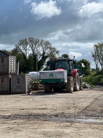 In Pike Corner, Ireland, a red tractor with a front-mounted planter attachment, labeled "UK PLANTER SOLUTIONS LIMITED," is parked on a dirt lot. The planter is silver and green. Several large wooden crates, some labeled "JOHN GABBIE & SONS PLANT RAISERS," are stacked to the left of the tractor. A large white cylindrical tank is visible behind the tractor. Trees and vegetation line the background, and the sky is partly cloudy. The ground is uneven and appears to be a mixture of dirt and gravel, with some puddles.