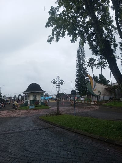A cloudy day scene features a paved road leading to an entrance. A small guardhouse with a domed roof sits to the left, while a building adorned with a stylized leaf-like sculpture is on the right. A decorative lamp post stands near a barrier across the road. Trees frame the top and right of the image, with palm trees visible in the background. The overall atmosphere is overcast and quiet.