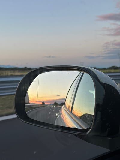 A side-view mirror of a vehicle reflects a sunset over a road. The sky in the reflection is a gradient of pale blue, pink, and orange, with faint clouds. The road ahead in the reflection appears to be an empty highway with a few car headlights visible in the distance. The exterior of the side-view mirror is black. The foreground of the image shows the dark gray car body. In the background outside the mirror, a guardrail runs along the side of the road, and a distant landscape of rolling hills and faint mountain ranges is visible under a pale sky. The location is the city of Malo Konare, Bulgaria.