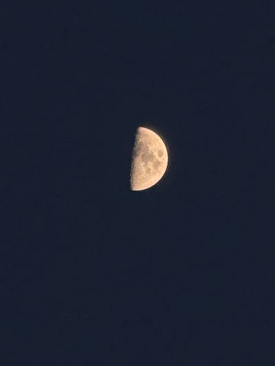 This image shows a close-up of the moon in a dark sky. The moon is partially illuminated, appearing as a crescent shape. The surface of the moon is visible, with craters and shadows indicating its texture. The sky is a deep, dark blue, suggesting it is nighttime. There are no other celestial bodies or atmospheric phenomena visible. The focus is entirely on the moon itself.