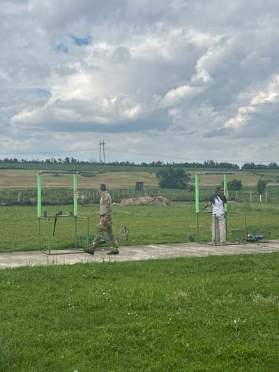 This is a medium shot of an outdoor clay pigeon shooting range, likely in or near the settlement of Feldmarshelsky, Russia, under a cloudy sky. Two individuals are present. On the left, a person in camouflage fatigues and a light-colored shirt walks away from a green shooting stand, which holds a rifle. On the right, a person wearing a white t-shirt with black sleeves and light-colored pants stands at a similar green shooting stand, holding a firearm and facing away from the camera. The ground is covered in green grass, with a concrete path leading to the shooting stands. In the background, there are fields, trees, and power lines, suggesting a rural or semi-rural environment. There are also piles of what appears to be hay or straw in the middle distance, and a wire basket containing targets or supplies near the right-hand shooting stand. The overall impression is of an active shooting sports venue.