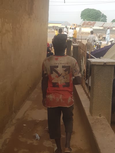 A street in Kufena, Nigeria, shows a man in a red and white "OFF-WHITE" shirt walking on a paved walkway. People are visible in the background on the street, alongside cars, mopeds, and structures. A large tree is in the distance.