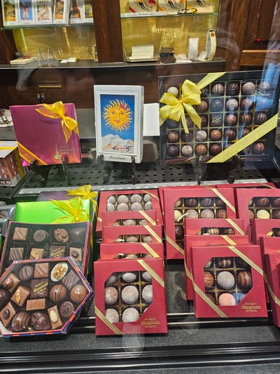 This image captures a display of assorted chocolates inside a confectionery shop. The chocolates are arranged in various boxes, some clear and some red, all adorned with yellow ribbons. The confectionery's name, "Confiserie Hanselmann St. Moritz," is visible on some of the packaging, indicating the location as St. Moritz, Switzerland. The display is set against a dark background, with a mirrored surface reflecting the scene. In the background, shelves are visible holding more items, possibly other confectioneries or related products. The lighting is bright, highlighting the chocolates and their packaging. There are no people visible in the image. The overall impression is of a well-organized and appealing product display.