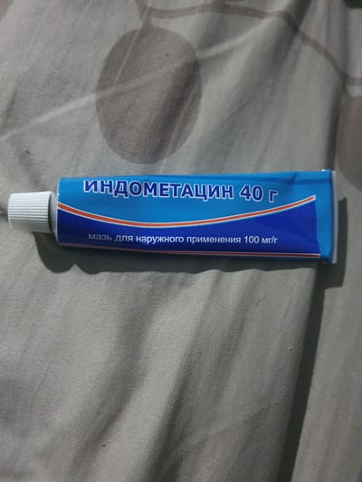 The image shows a close-up, angled shot of a blue and white tube of ointment, possibly for topical application, resting on a gray, textured fabric surface. The tube has white text in Cyrillic script that reads "ИНДОМЕТАЦИН 40 г" (Indometacin 40 g) and below that, "мазь для наружного применения 100 мг/г" (ointment for external use 100 mg/g). The cap of the tube is white and ribbed. The fabric surface appears to be a blanket or bedspread, with subtle shadowing and wrinkles, suggesting it is a domestic or personal setting. There is no indication of the city of Ghafurov, Tajikistan within the image itself.