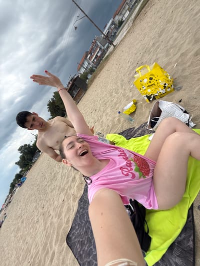 The image depicts two individuals at a beach. One person is lying on a beach towel and wearing a pink shirt with a "Raspberry" design. They are holding the camera at arm's length. The second individual is standing behind the first person. The beach sand is light colored, and a yellow shopping bag, a white bag, and a water bottle are scattered nearby. Buildings and overcast skies are visible in the background, suggesting a location in Νέα Φλογητά, Greece.