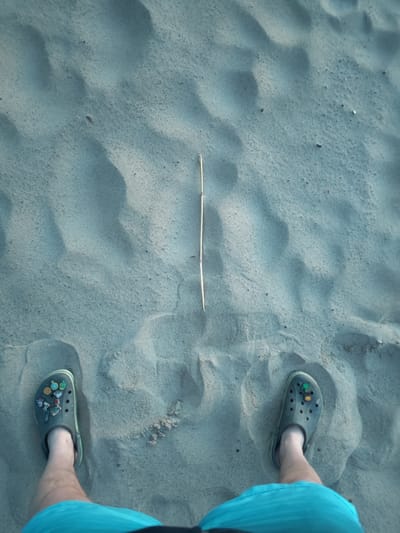 In the city of Motornoe, Russia, an individual stands on a sandy surface, viewed from a top-down perspective. The sand exhibits undulating patterns, indicating the presence of dunes or ripples. The individual is wearing two dark-colored crocs, each adorned with various colorful charms. On the left croc, charms depicting various small objects like a globe, a small building, a person figure, and a red bus are visible. The right croc features a green charm and a small yellow and red charm. The individual is also wearing turquoise shorts. Centrally positioned between the person's feet is a thin, light-colored stick lying horizontally on the sand. The lighting suggests it is daytime, with shadows cast by the sand's contours.